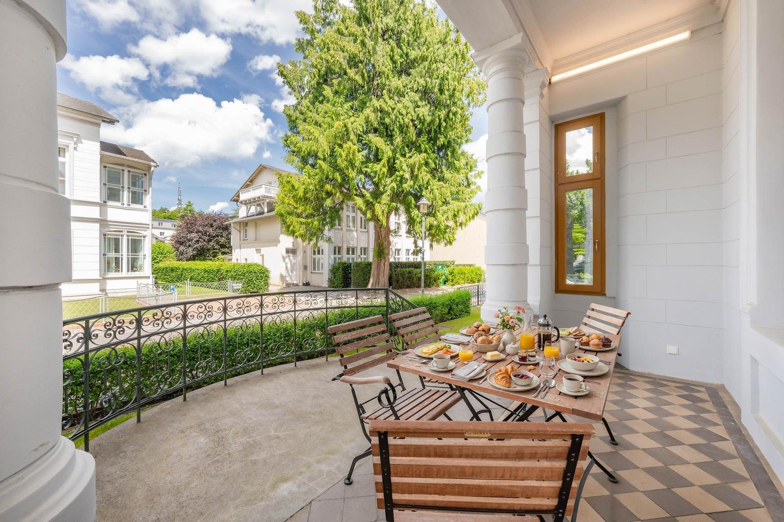 Terrasse mit Tisch, Stühlen und Frühstück. Blick auf grüne Bäume und weiße Gebäude.