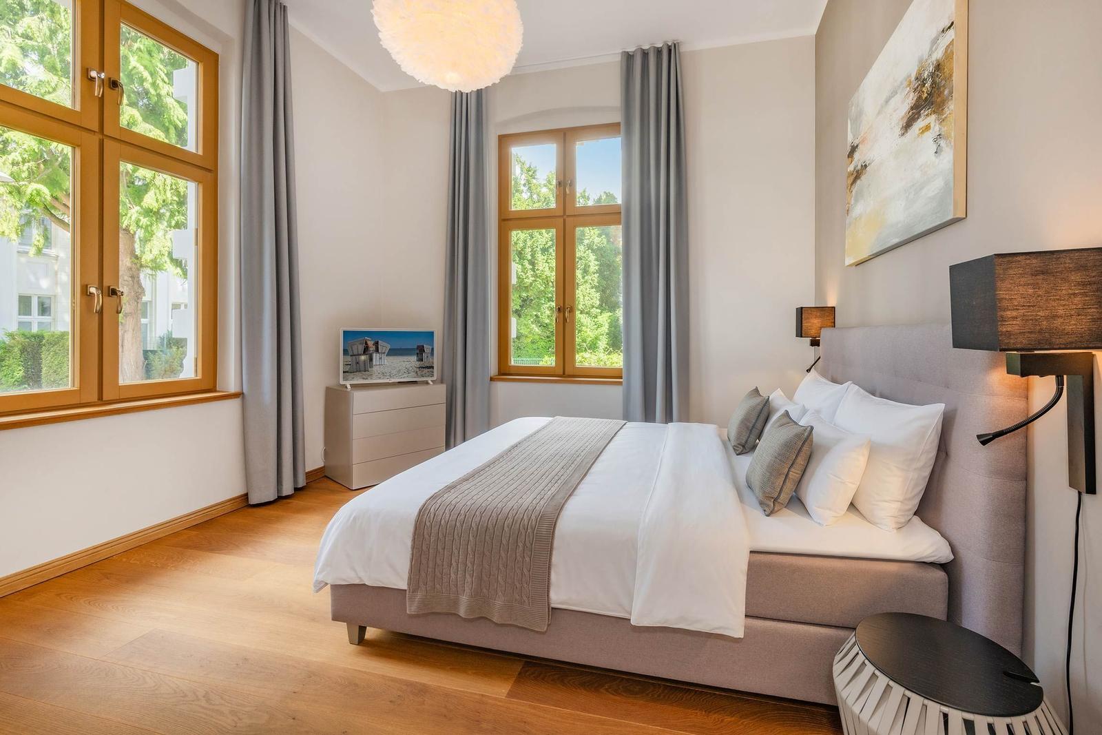 Schlafzimmer mit großem Bett, Holzboden und zwei Fenstern mit Blick auf Grün.