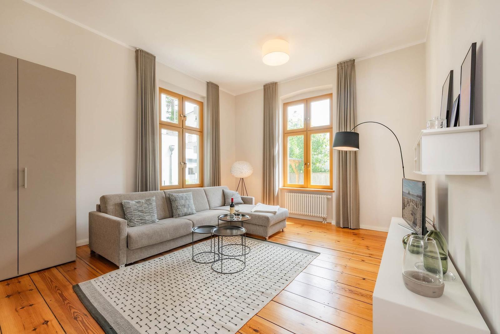 Wohnzimmer mit Sofa, zwei Fenstern, Holzboden und TV-Schrank.