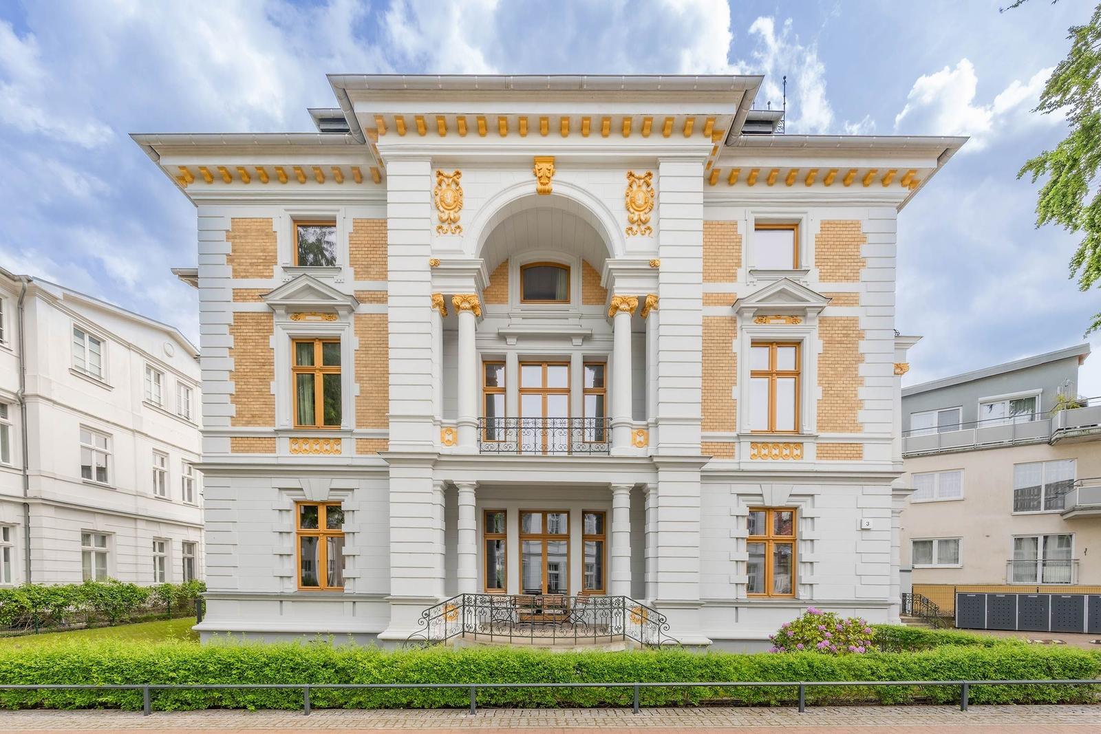 Haus mit weißer Fassade, goldfarbenen Verzierungen und Holzfenstern. Vorgarten mit grüner Hecke.
