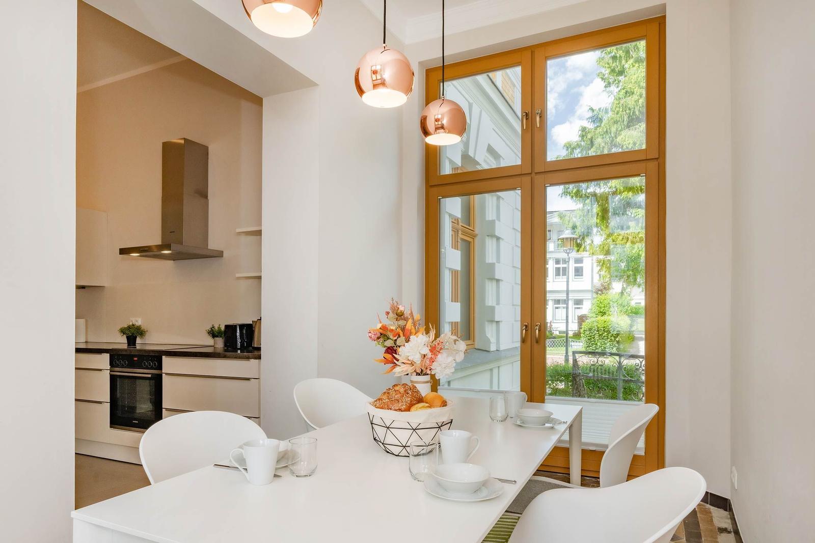 Essbereich mit weißem Tisch, vier Stühlen und Blick auf Balkon. Küche im Hintergrund.