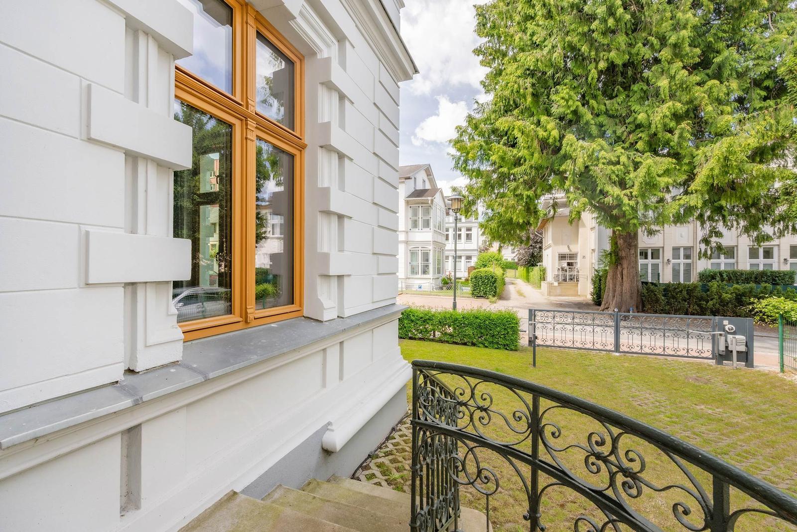 Haus mit weißer Fassade, Holzfenster und schmiedeeisernem Geländer. Grünanlage und Bäume im Hintergrund.