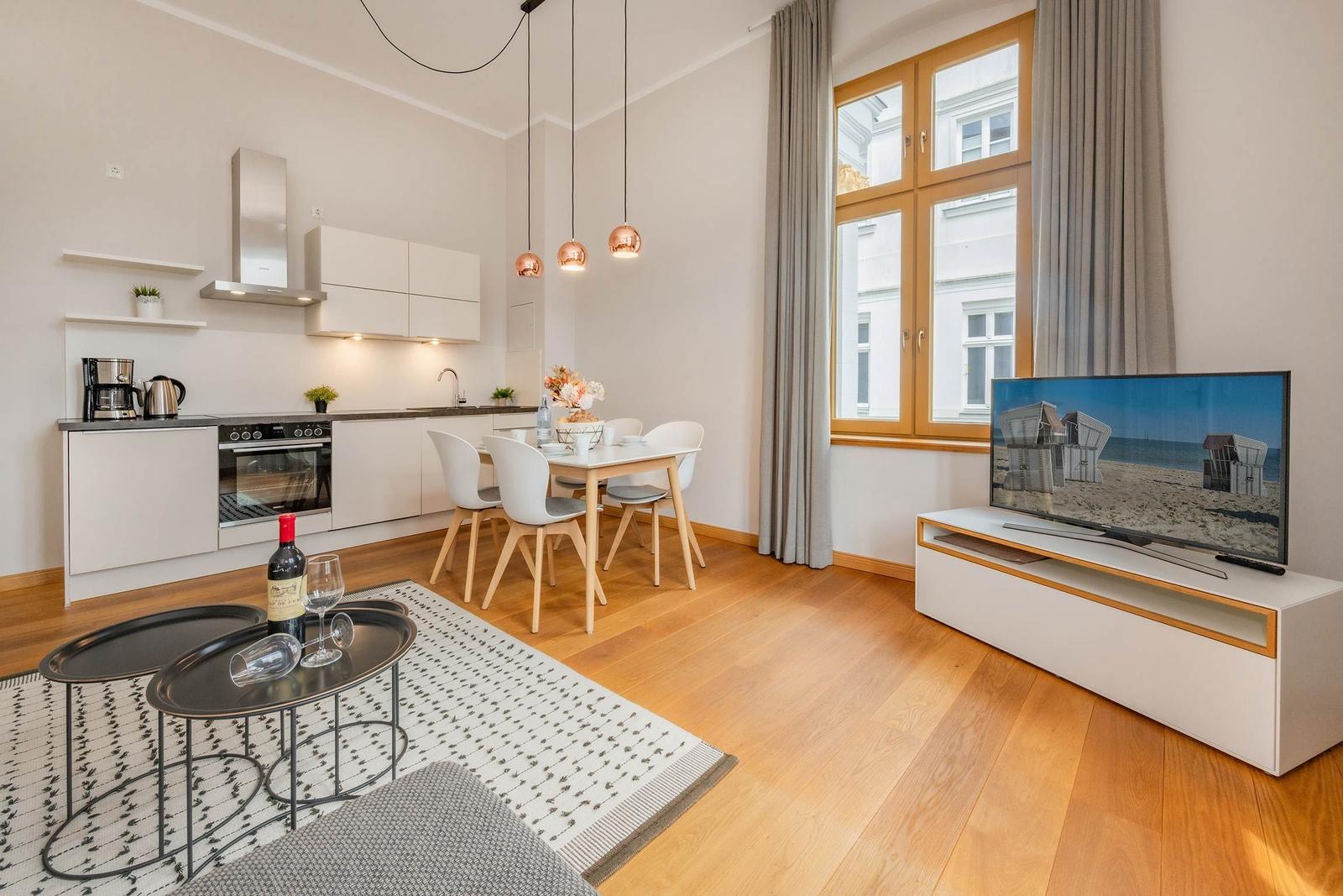 Wohnung mit Küche, Esstisch und Fernseher. Holzboden und großes Fenster.