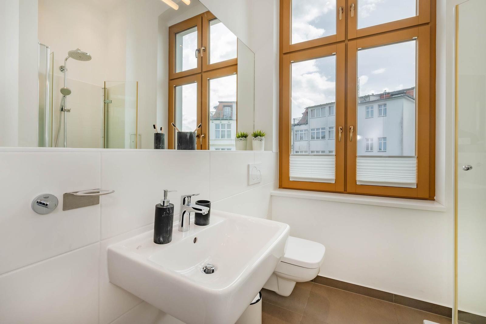 Badezimmer mit Waschbecken, Toilette und Dusche. Große Fenster mit Holzrahmen.