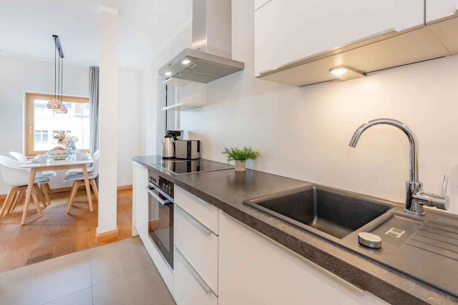 Moderne Küche mit Arbeitsplatte, Spüle und Dunstabzug. Im Hintergrund ist Essbereich mit Tisch und Stühlen.