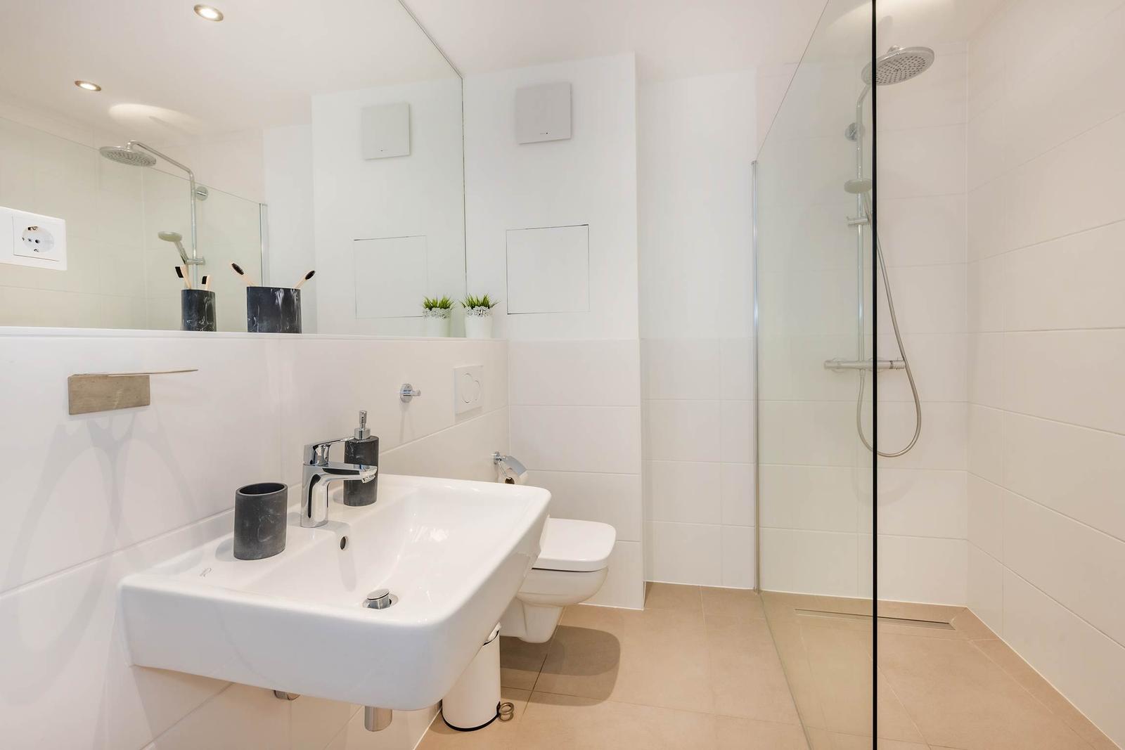 Badezimmer mit Dusche, Waschbecken und Toilette. Weiße Fliesen und Spiegel.