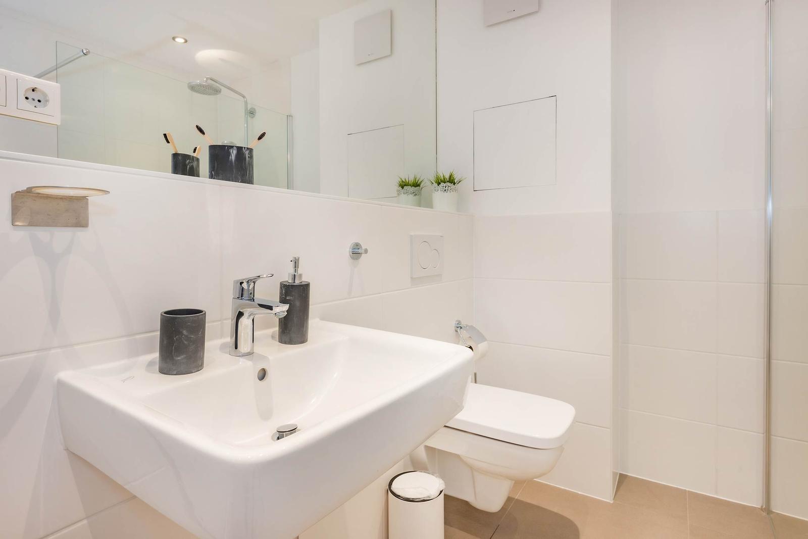 Badezimmer mit Waschbecken, Toilette und Dusche. Weiße Fliesen und Spiegel.