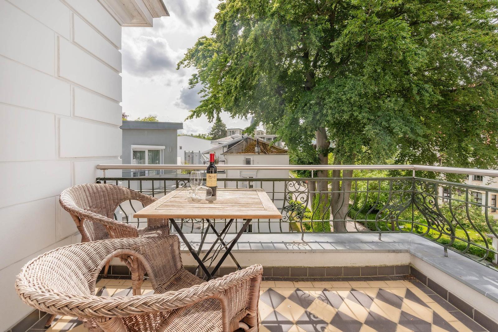 Balcony with wicker chairs, table, and wine bottle. View of trees and buildings.