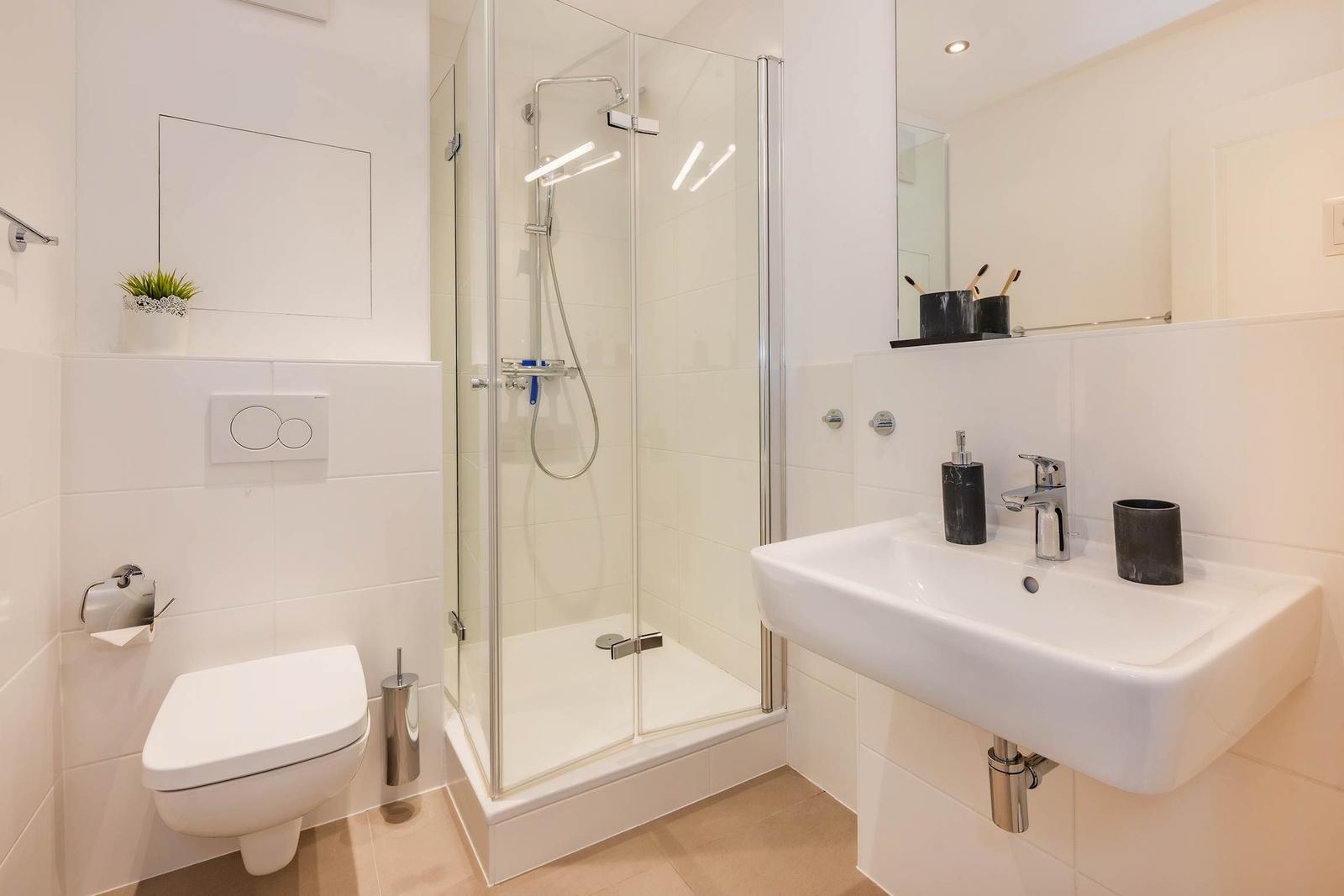 Badezimmer mit Dusche, Waschbecken und Toilette. Weiße Fliesen und moderne Armaturen.