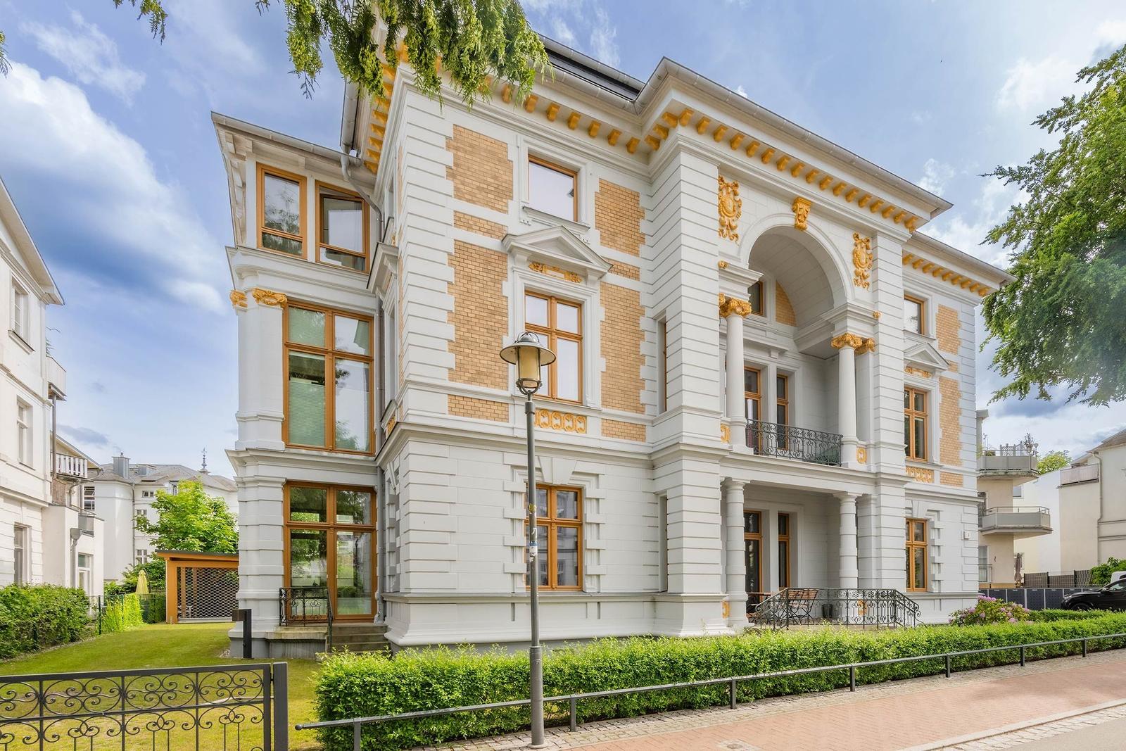 Elegantes Haus mit weißen Wänden, gelben Ziegelsteinen und goldenen Verzierungen. Große Fenster und Balkon.