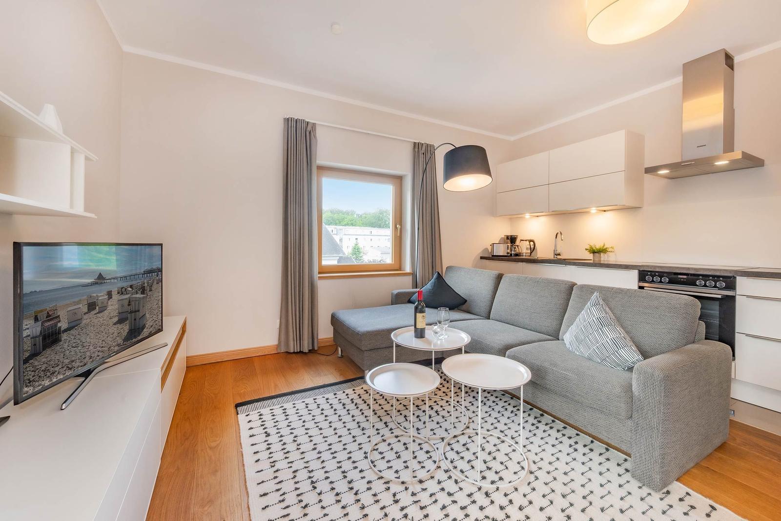 Wohnzimmer mit Sofa, Fernseher und Küchenzeile. Fenster mit Blick auf Außengebiet.