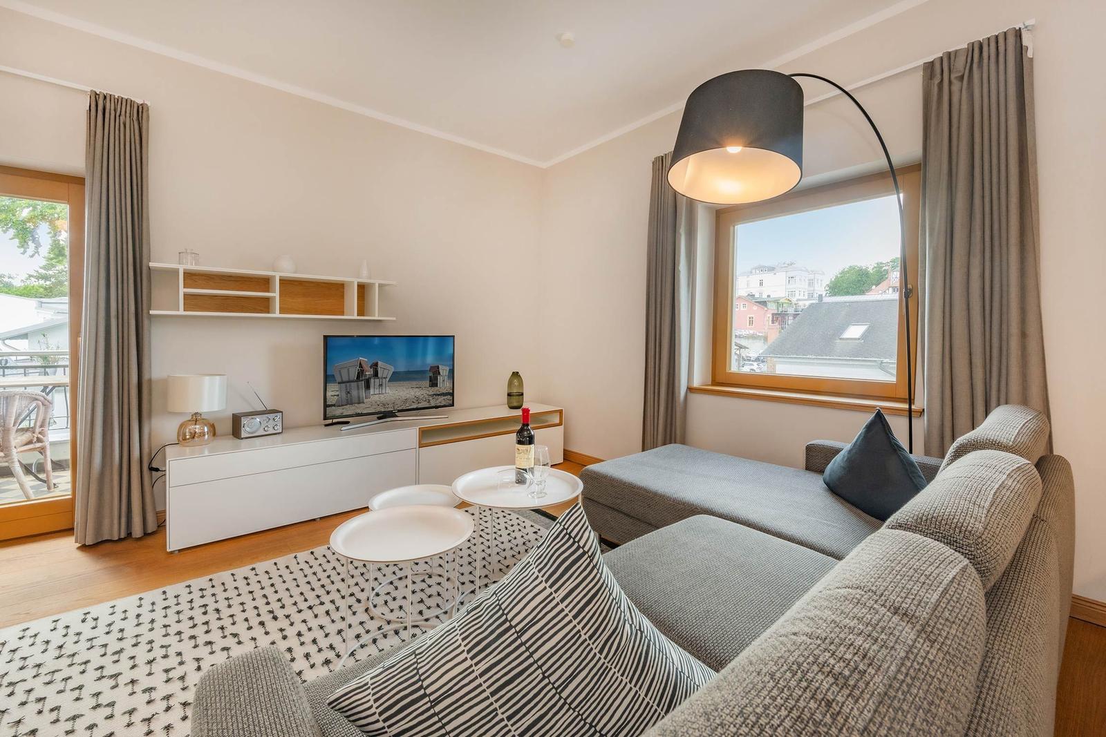 Wohnzimmer mit Sofa, TV und Fensterblick. Es gibt einen Tisch und eine Stehlampe.