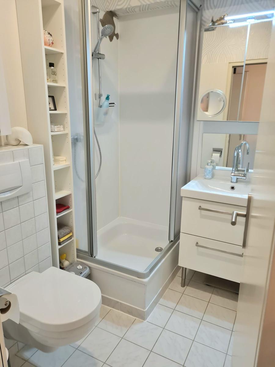 Badezimmer mit Dusche, Waschbecken und Toilette. Weiße Fliesen und Regale.