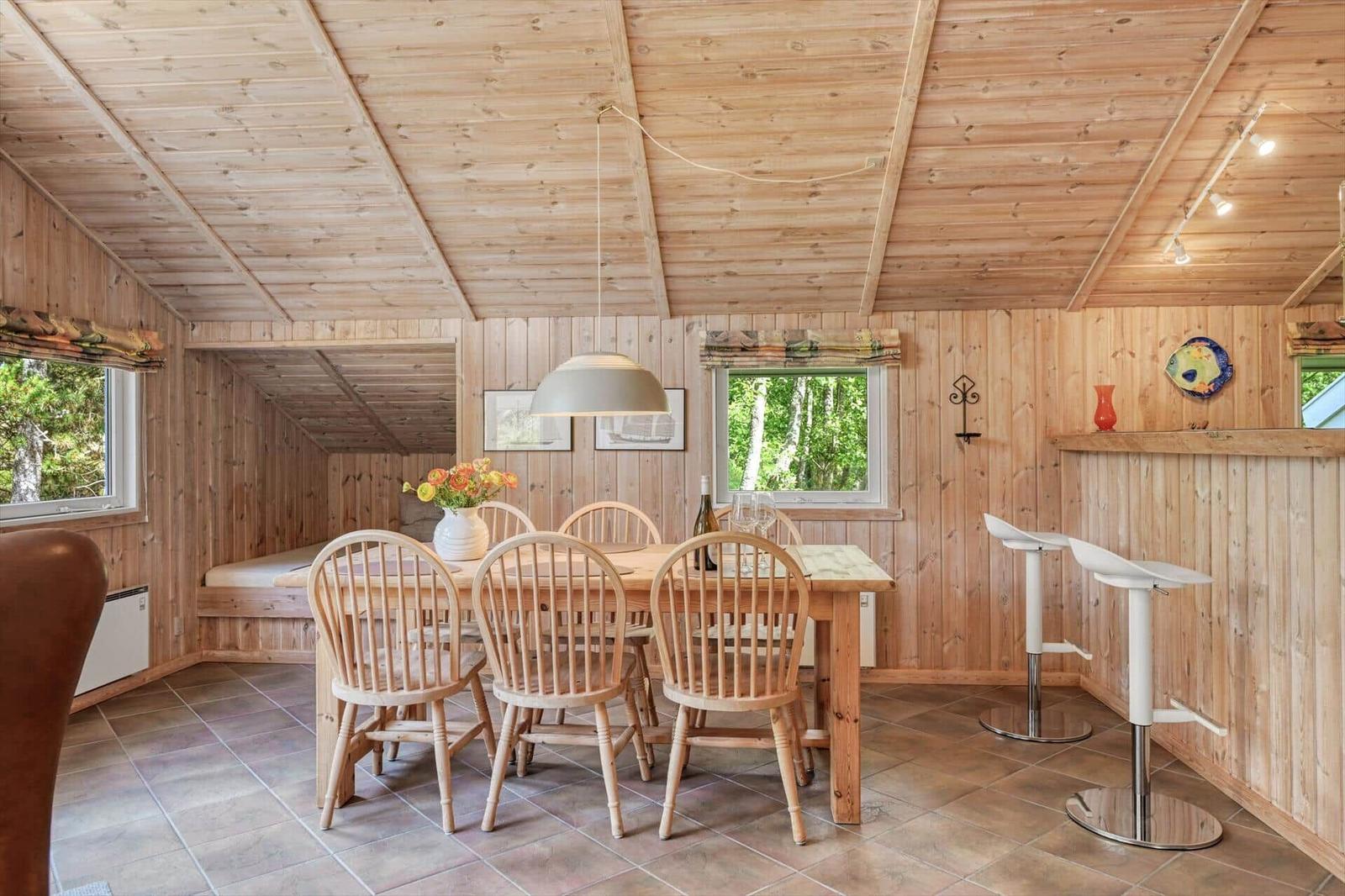 Essbereich mit Holztisch, Stühlen und Fenstern. Holzwände und -decke. Barhocker an der Wand.