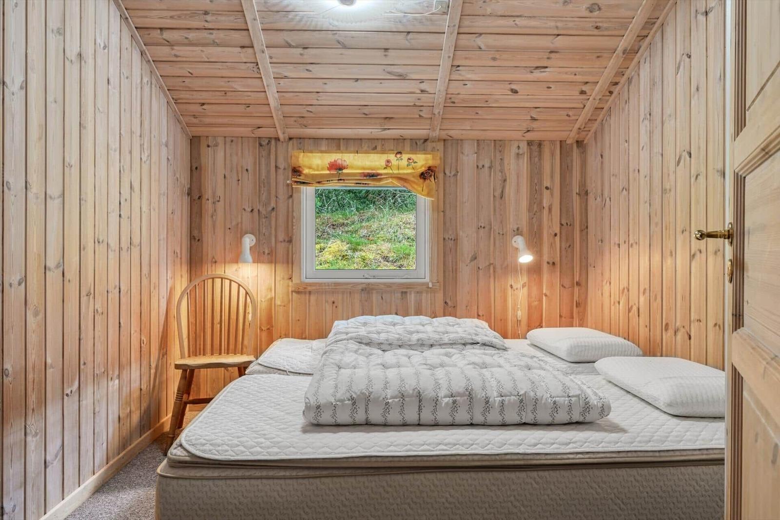 Schlafzimmer mit Holzwänden, Bett, Stuhl und Fenster mit Blick auf Grün.