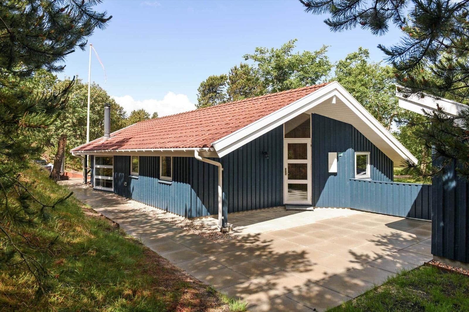 Ein blau gestrichenes Haus mit rotem Dach und weißer Tür. Umgeben von Bäumen und einem gepflasterten Weg.