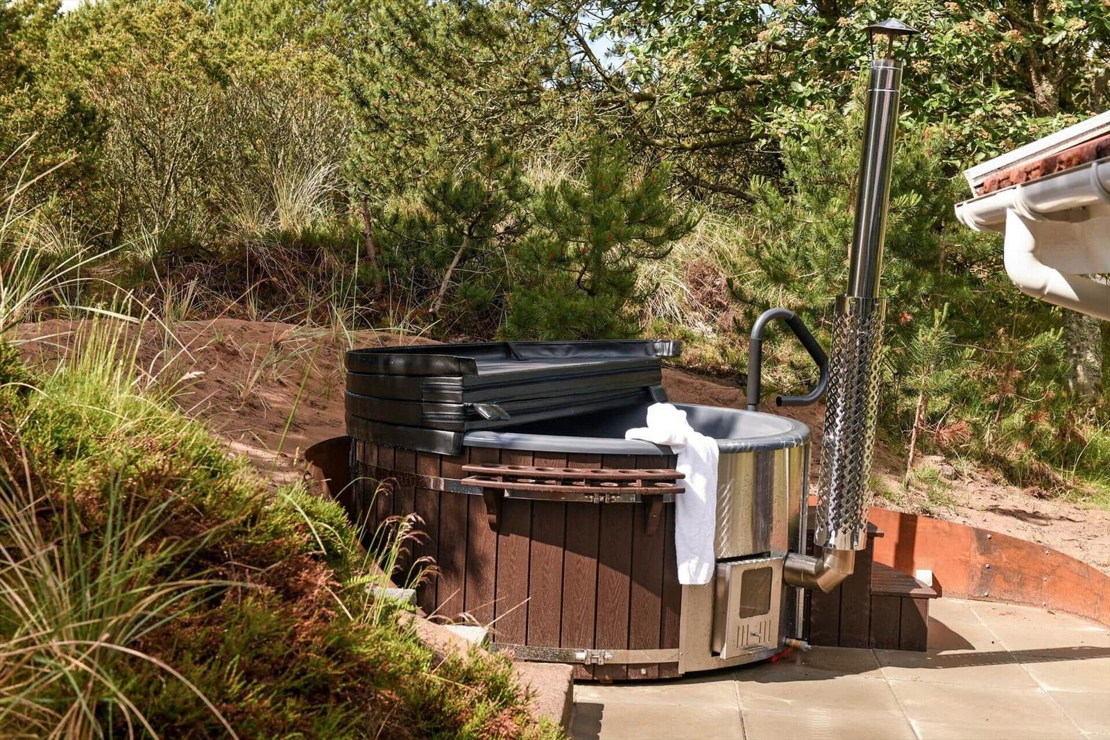 Außenwhirlpool mit Holzrahmen und Metallrohr, umgeben von Grünflächen und Bäumen.
