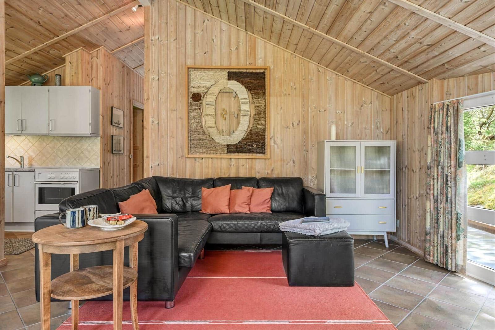 Wohnzimmer mit schwarzer Couch, rotem Teppich und Holzwand. Küche im Hintergrund.