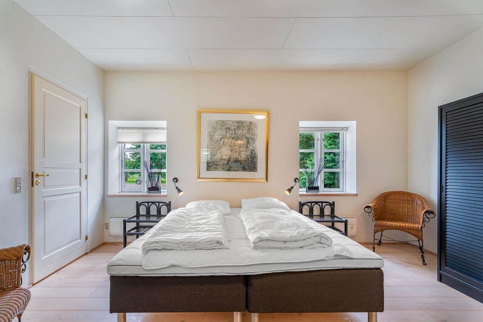 Schlafzimmer mit Bett, zwei Fenstern, Wandbild und Stuhl.