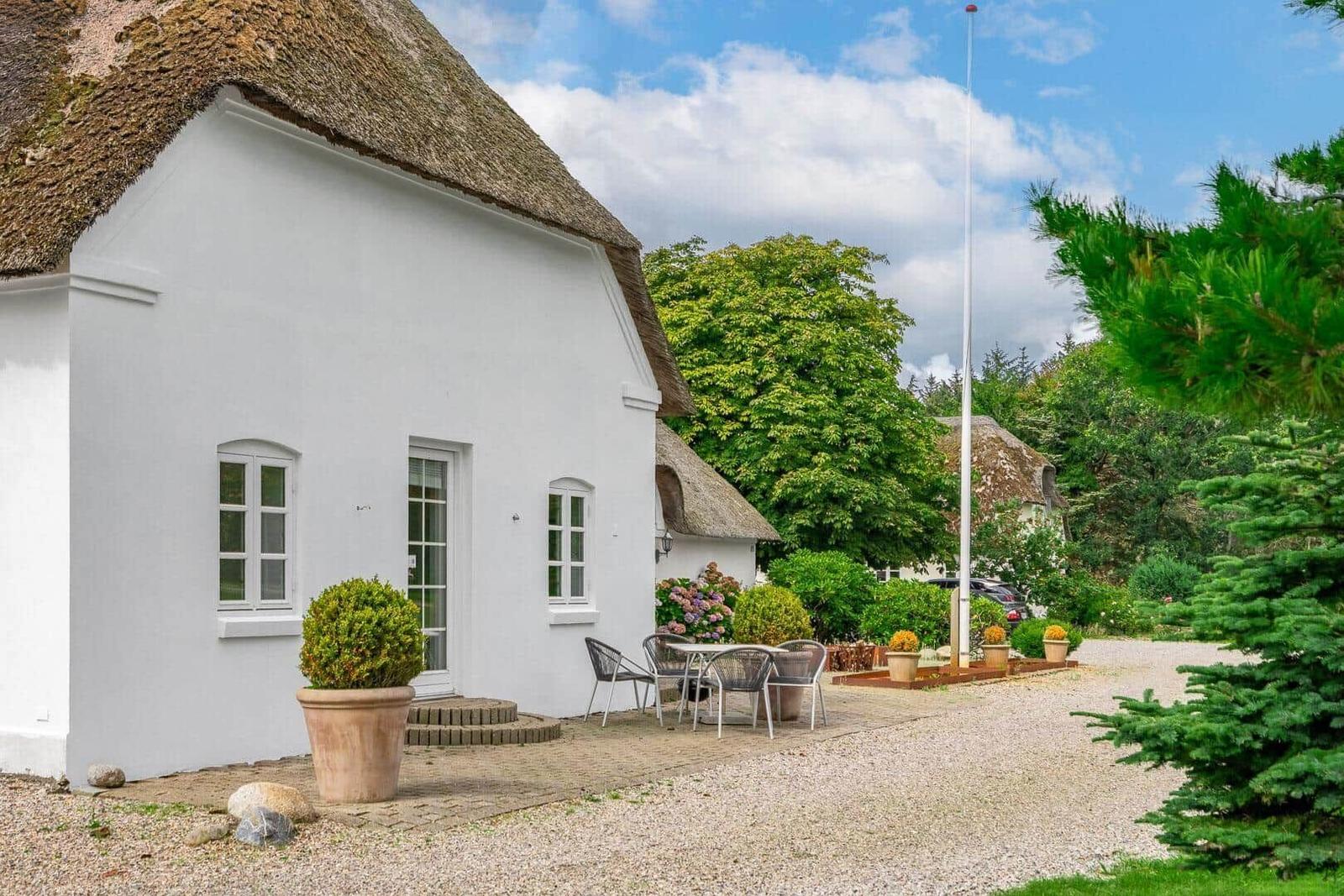 Weißes Haus mit Strohdach, Terrasse und Garten. Holztisch, Stühle, Pflanzen.