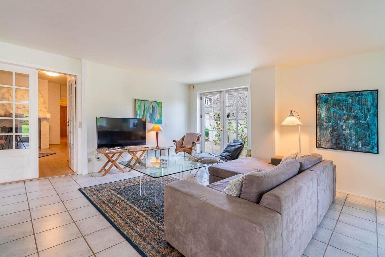 Wohnzimmer mit Sofa, Glas-Tisch und Fernseher. Durchgänge zu anderen Räumen und Fenster mit Blick nach draußen.