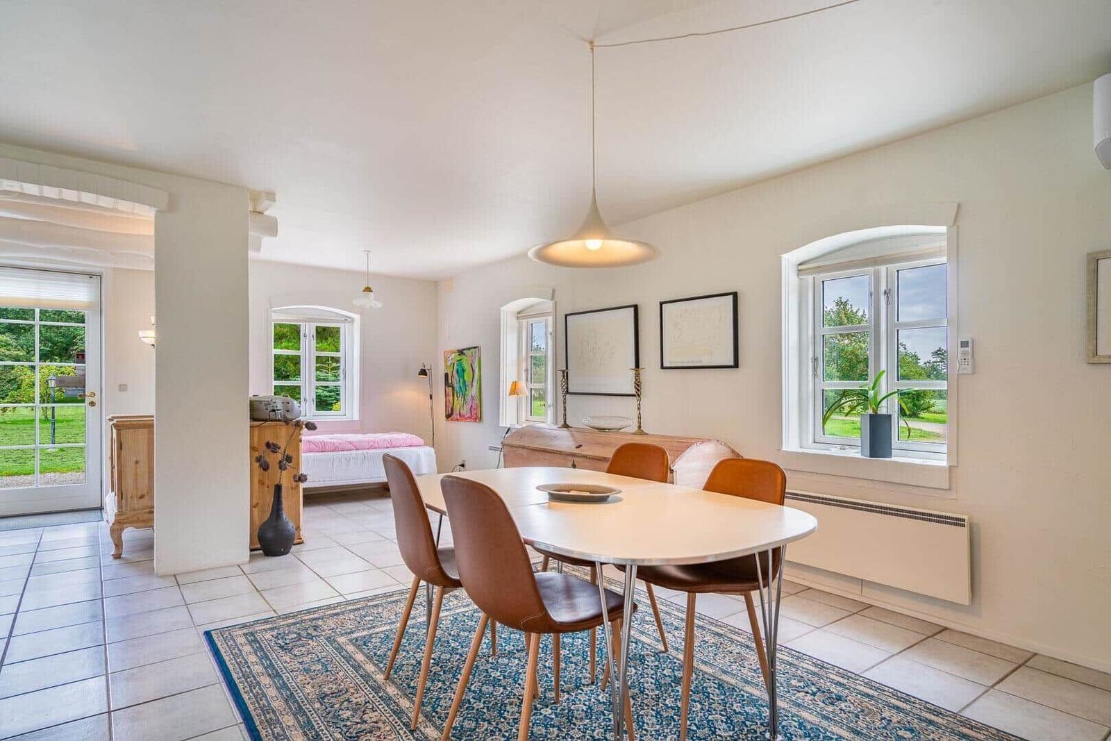Essbereich mit Tisch und Stühlen auf Teppich. Im Hintergrund Schlafzimmer und Fenster mit Blick auf Garten.