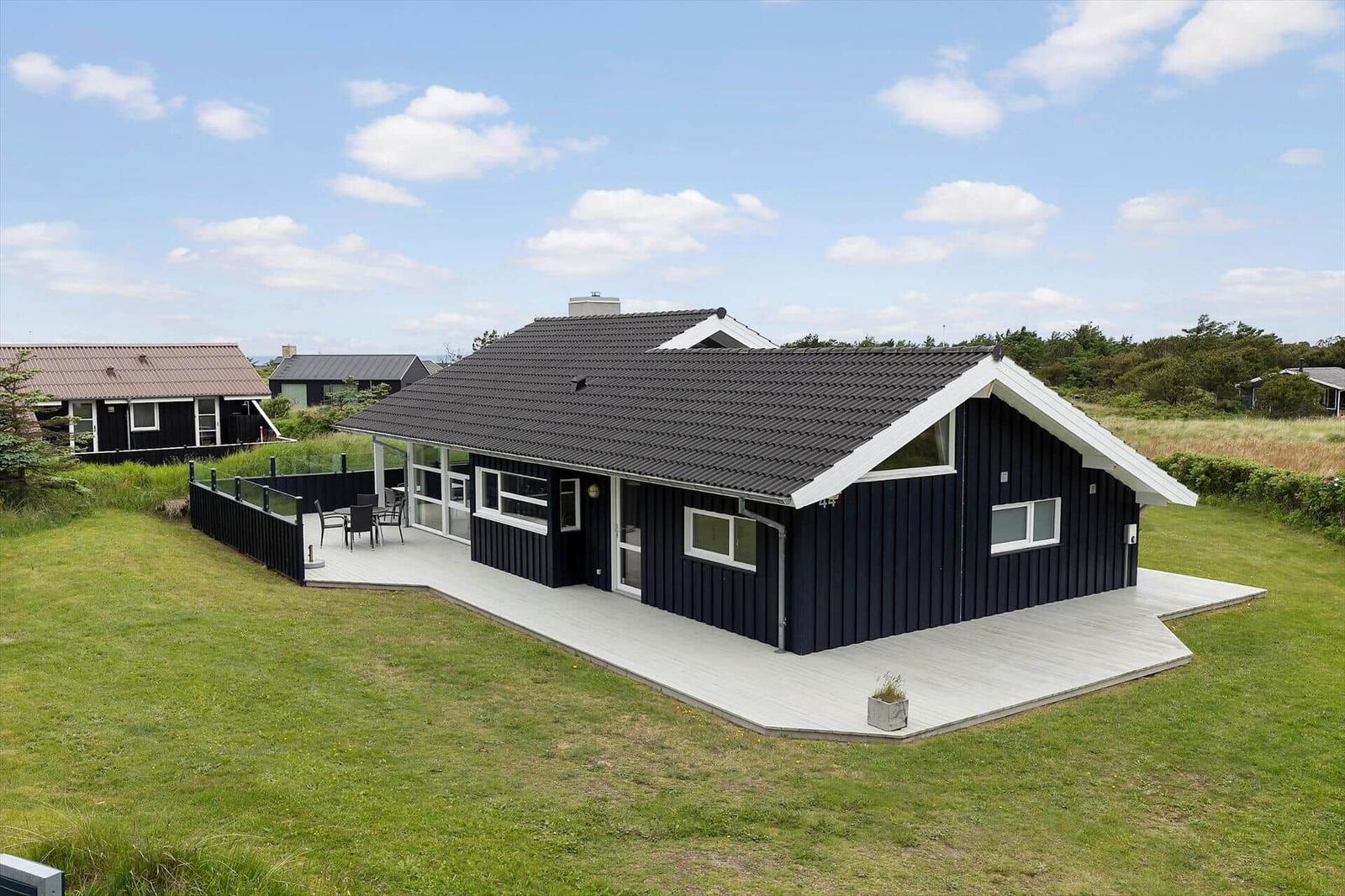 Ein schwarzes Ferienhaus mit weißem Holzdeck und Garten. Hintergrund: Gras und Bäume.