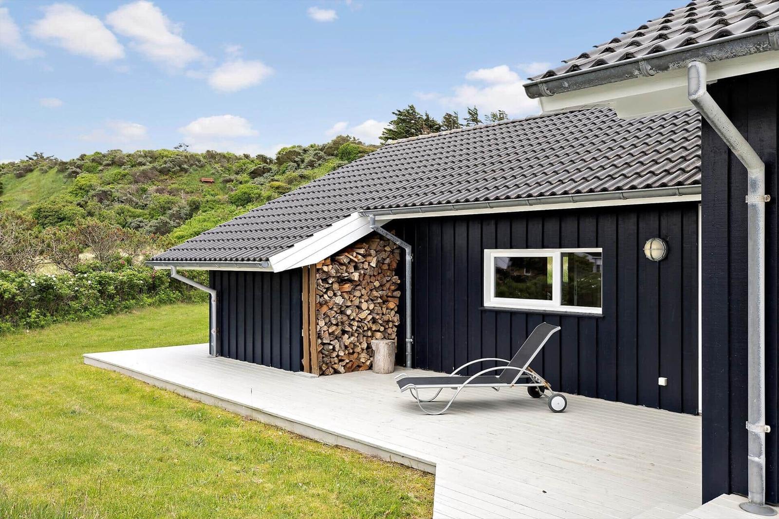 Schwarzes Haus mit Holzstapel, Liegestuhl und Terrasse vor grüner Landschaft.