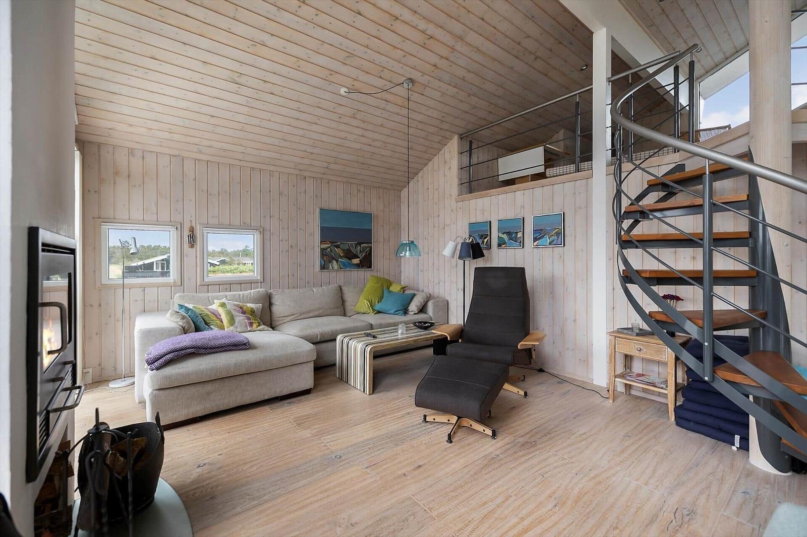 Wohnzimmer mit Sofa, Sessel und Spiralrampe. Holzwände und Boden. Kamin und Fenster.