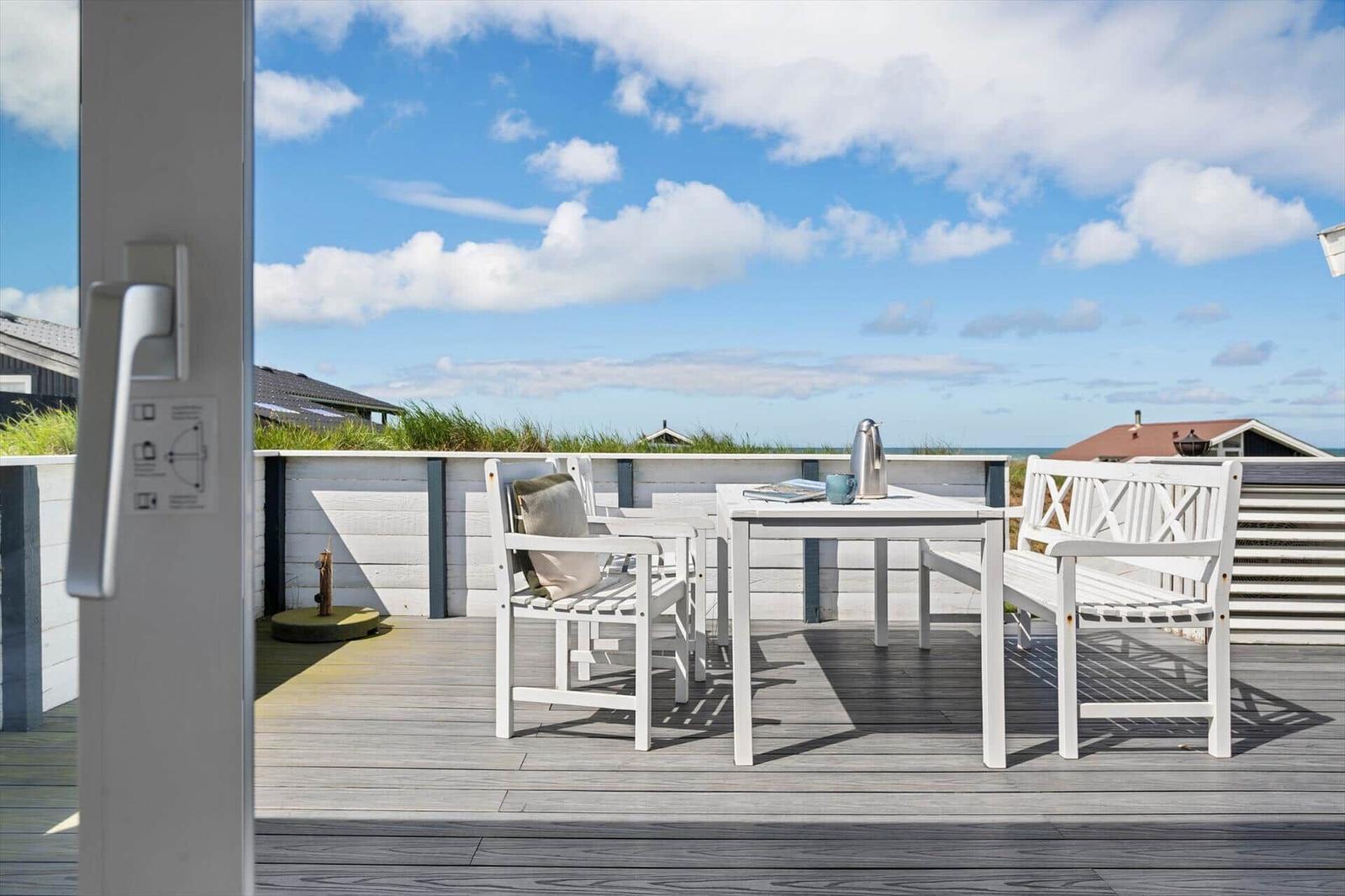 Terrasse mit weißem Tisch und Stühlen, Blick auf Strand und Himmel.