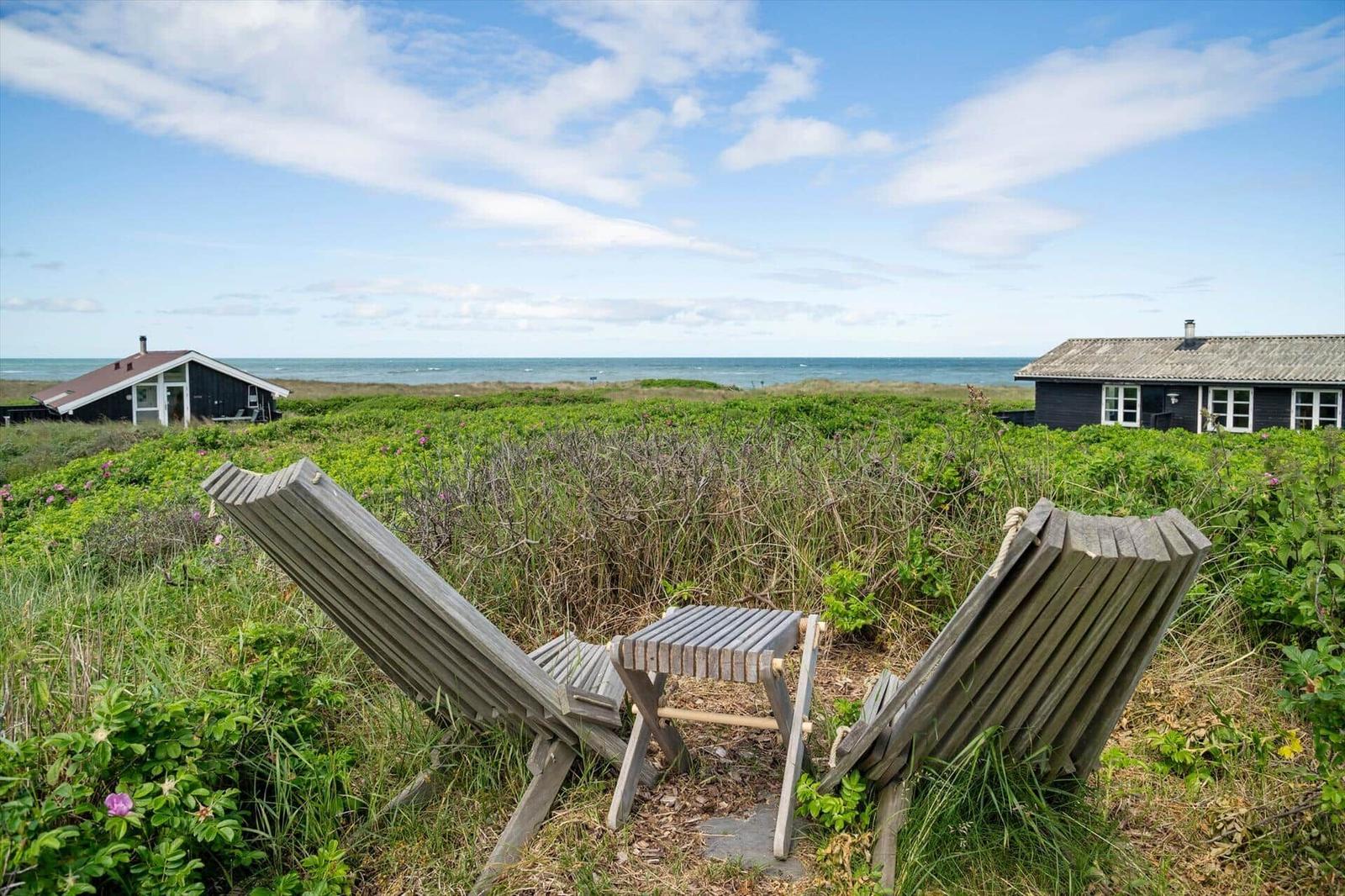 Zwei Holzliegestühle und ein Tisch im Gras mit Blick auf das Meer und Häuser.