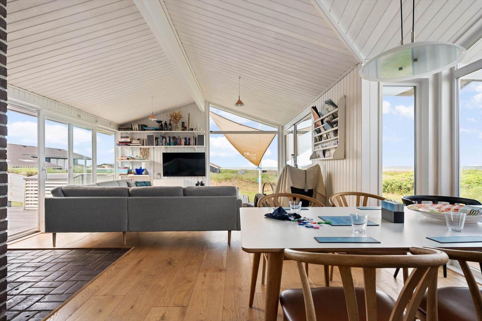 Wohn- und Essbereich mit Sofa, Tisch und Stühlen. Große Fenster mit Blick auf die Landschaft.