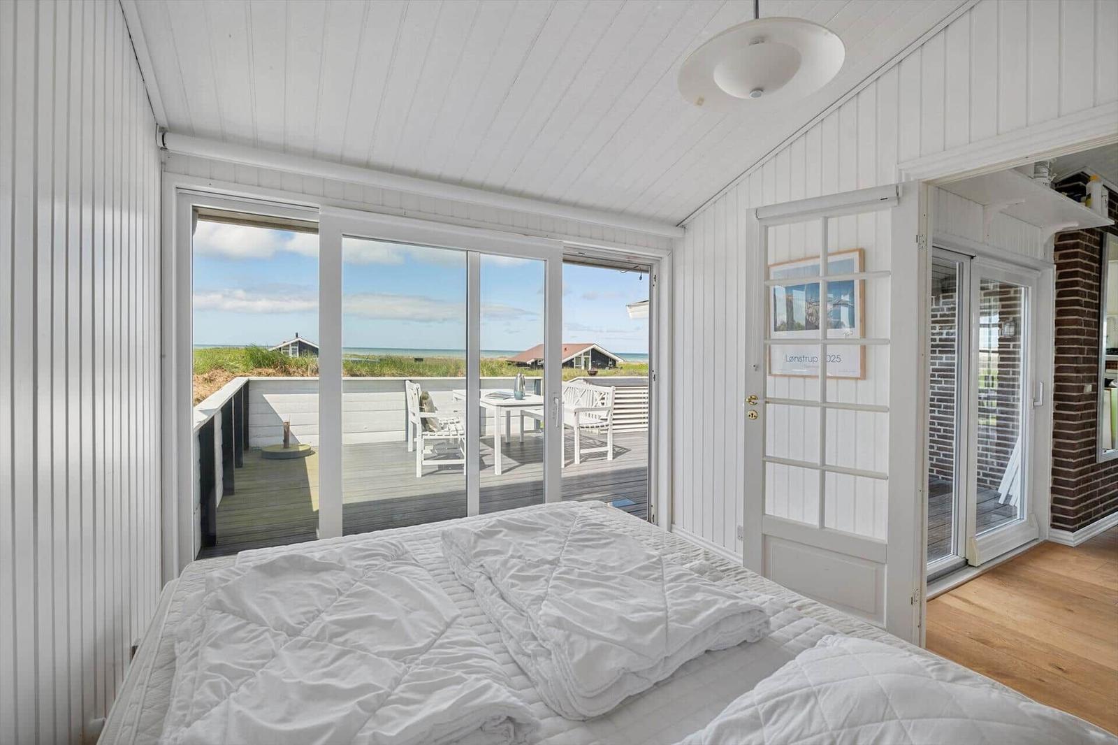Schlafzimmer mit Blick auf Terrasse und Strand. Weißes Bett, weiße Wände und Holzboden.