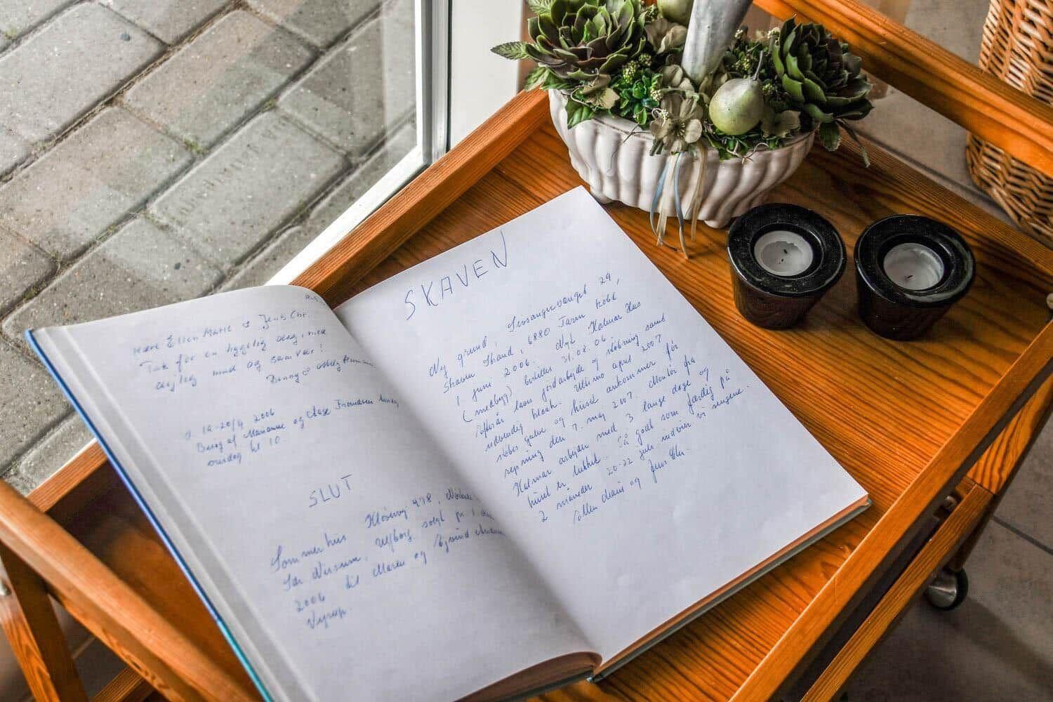 Ein Buch mit handschriftlichen Einträgen liegt auf einem Holztisch neben einer Pflanze und Kerzen.