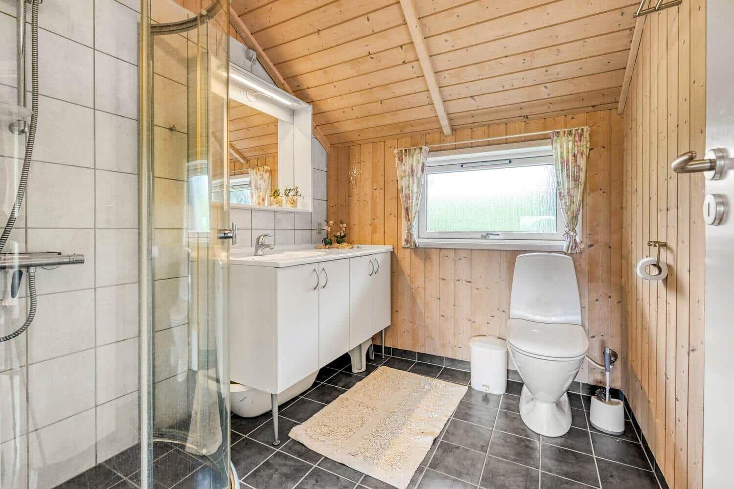 Badezimmer mit Dusche, Waschbecken und Toilette. Holzwände und Fenster mit Vorhängen.