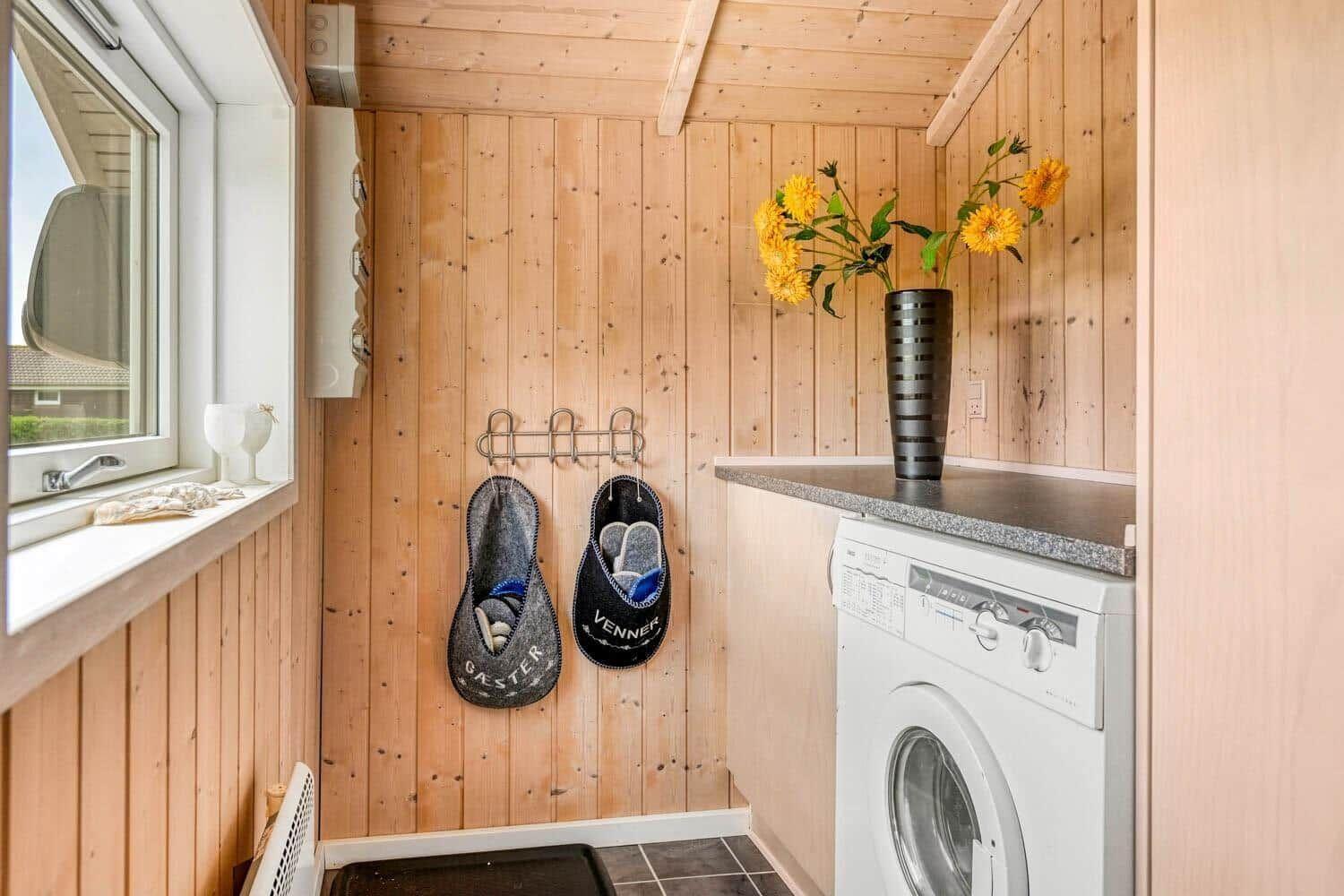 Wohnraum mit Waschmaschine, Holzwand und Fenster. Auf der Arbeitsplatte steht eine Vase mit Sonnenblumen.