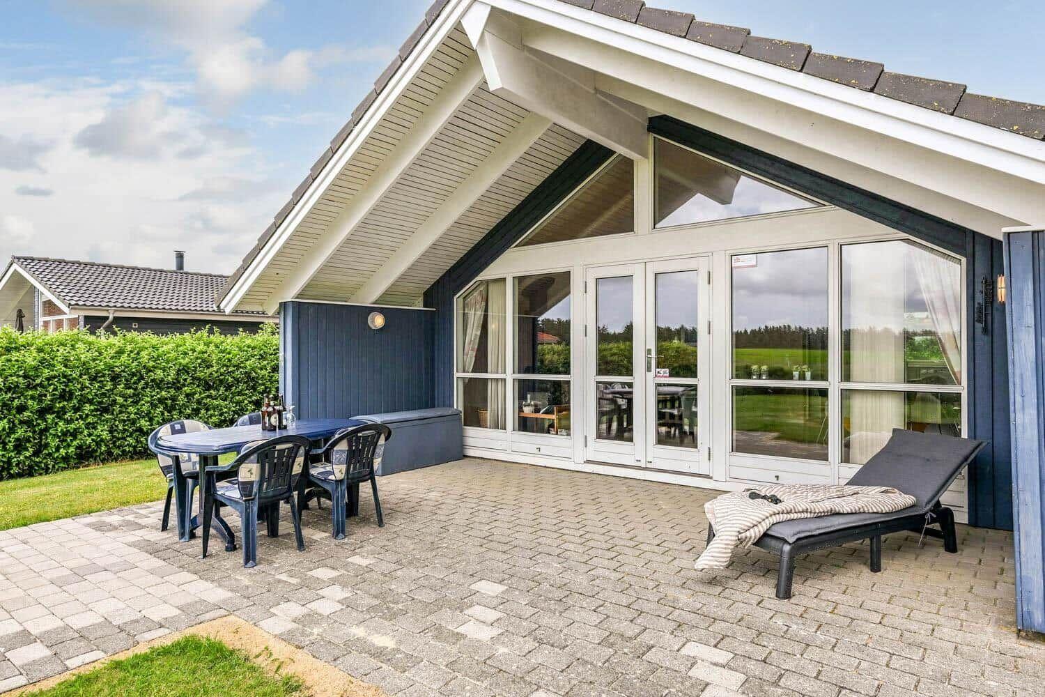 Terrasse mit Tisch, Stühlen und Liegestuhl vor einem Haus mit Glasfront.