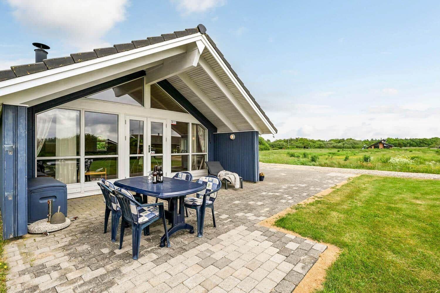 Haus mit Terrasse, Tisch und Stühlen. Grünflächen und Wald im Hintergrund.