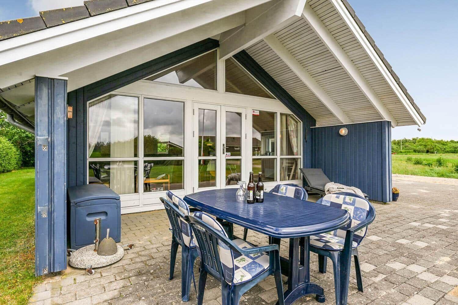 Terrasse mit blauem Tisch und Stühlen, Glasfront und Grill. Grün hinter dem Haus.