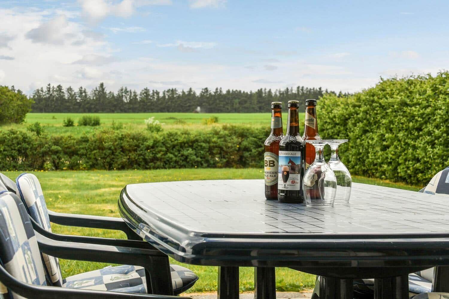 Außenbereich mit Tisch, Stühlen und Bierflaschen vor grünem Feld und Hecken.