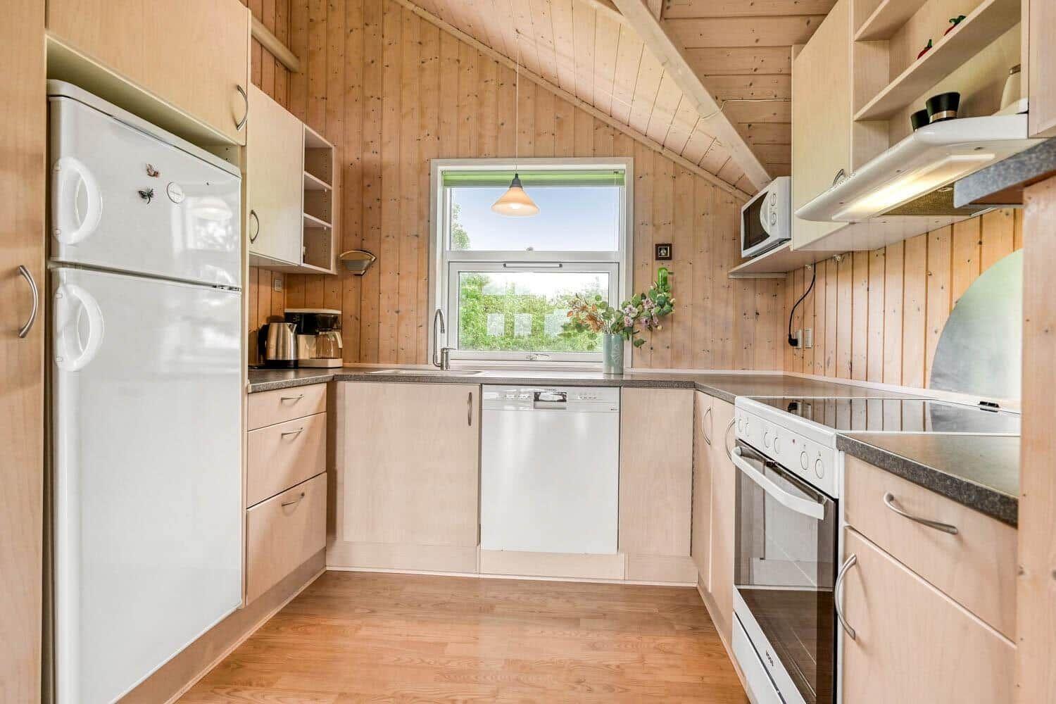 Küche mit Holzwänden, weißer Kühlschrank, Spülmaschine und Fenster mit Blick nach draußen.