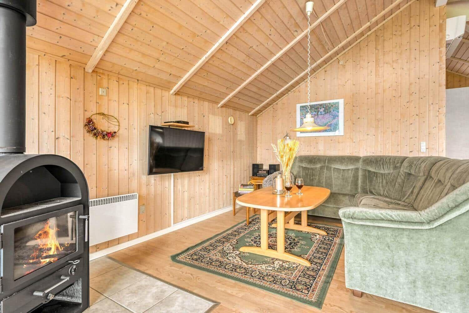 Wohnraum mit Holzwand, Sofa, Tisch und Kaminofen mit brennendem Feuer.