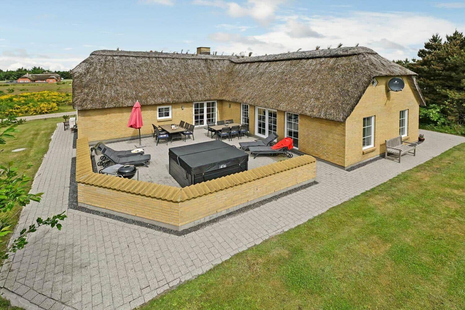 Haus mit Strohdach, Terrasse mit Liegen und Grillplatz, umgeben von Grünfläche.