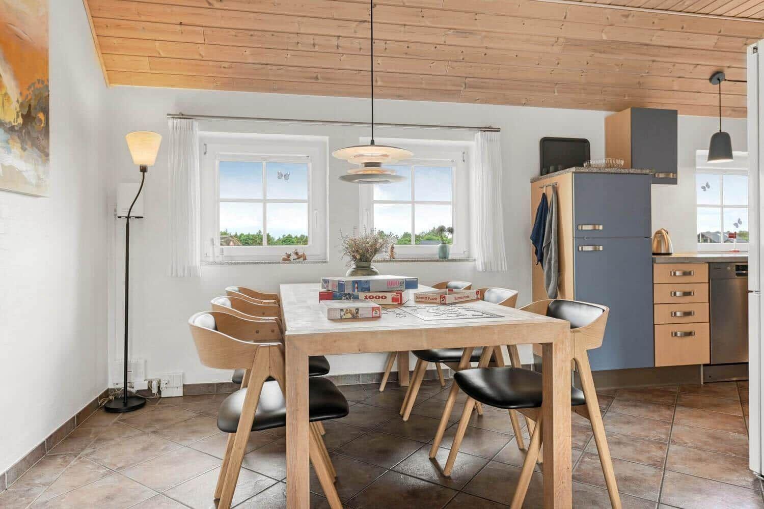 Esszimmer mit Holztisch, Stühlen und Küchenbereich. Holzdecke und Fenster mit Blick.