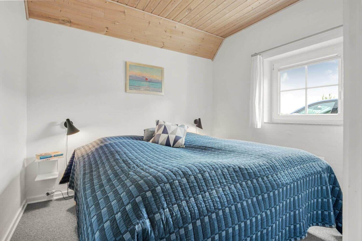 Schlafzimmer mit Bett, blauer Decke, Fenster und Holzdecke.