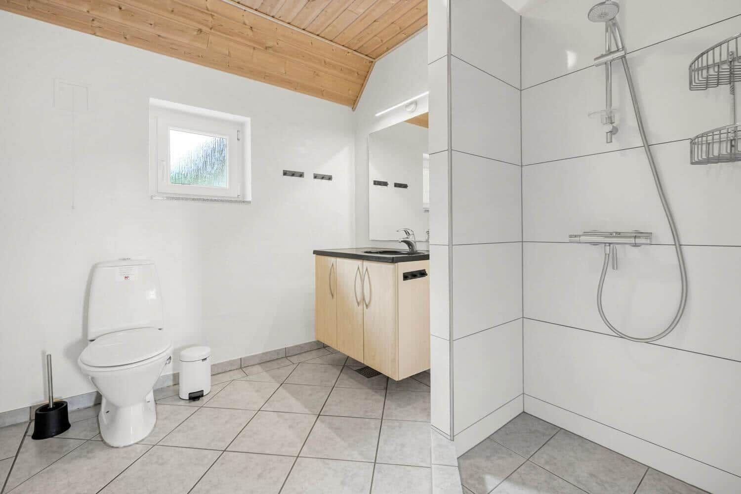 Badezimmer mit Dusche, Waschbecken und Toilette. Holzdecke und Fenster sichtbar.