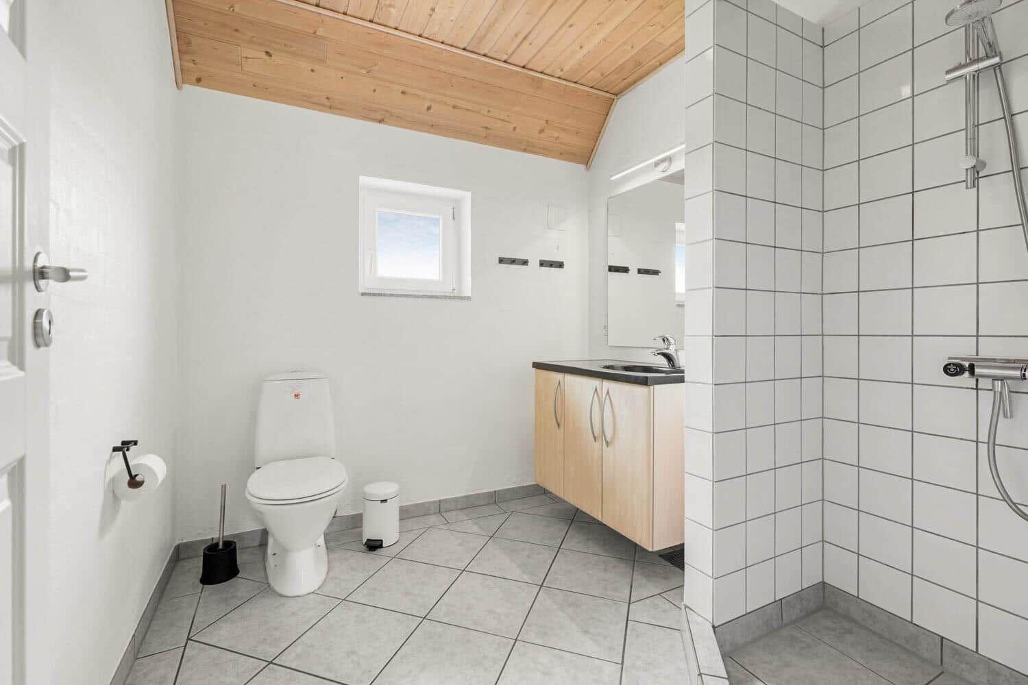 Badezimmer mit Toilette, Waschbecken und Dusche. Holzdecke und weiße Fliesen.