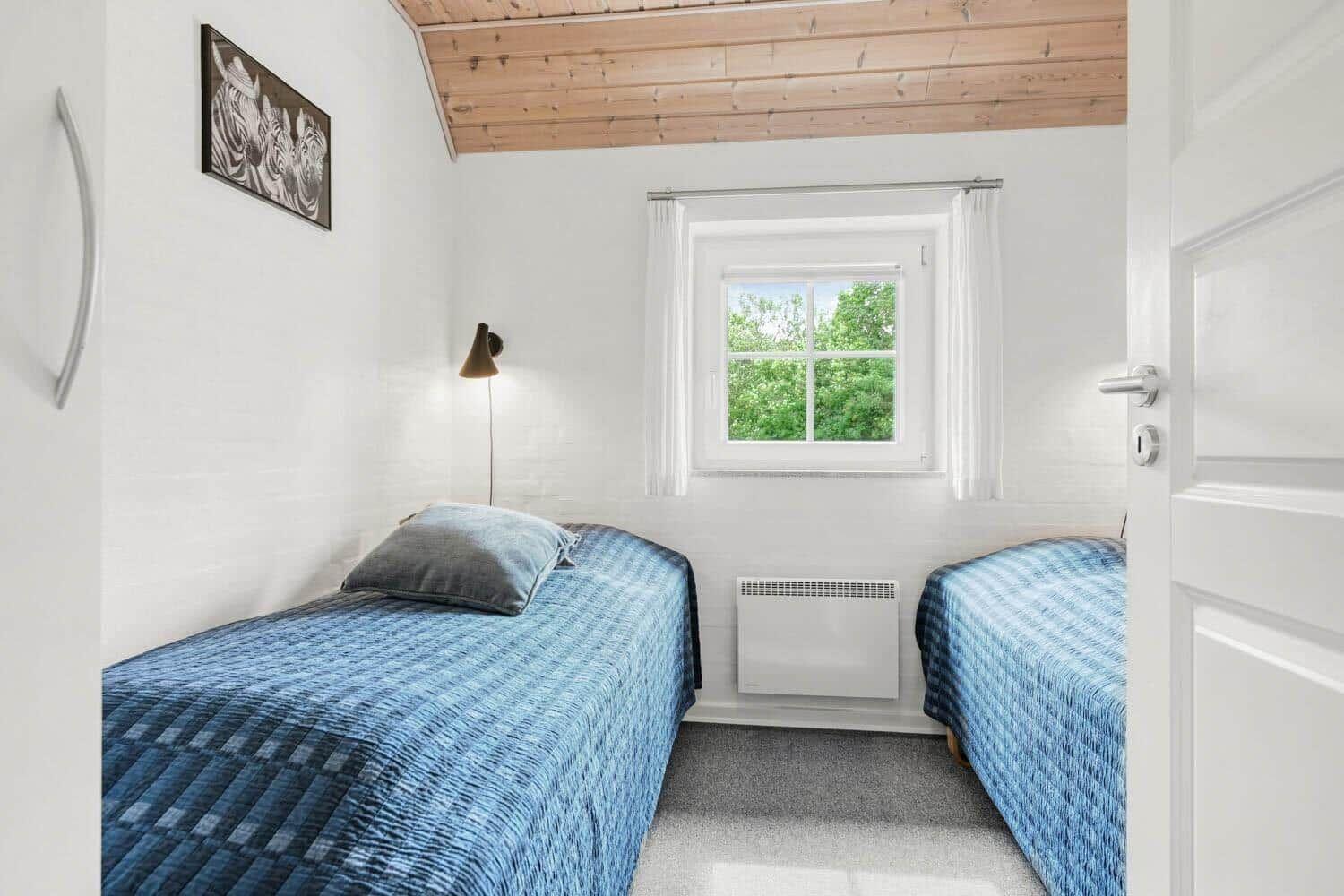 Zwei Einzelbetten mit blauen Bettwäschen, weißer Wand und Holzdecke. Fenster mit Blick auf Bäume.