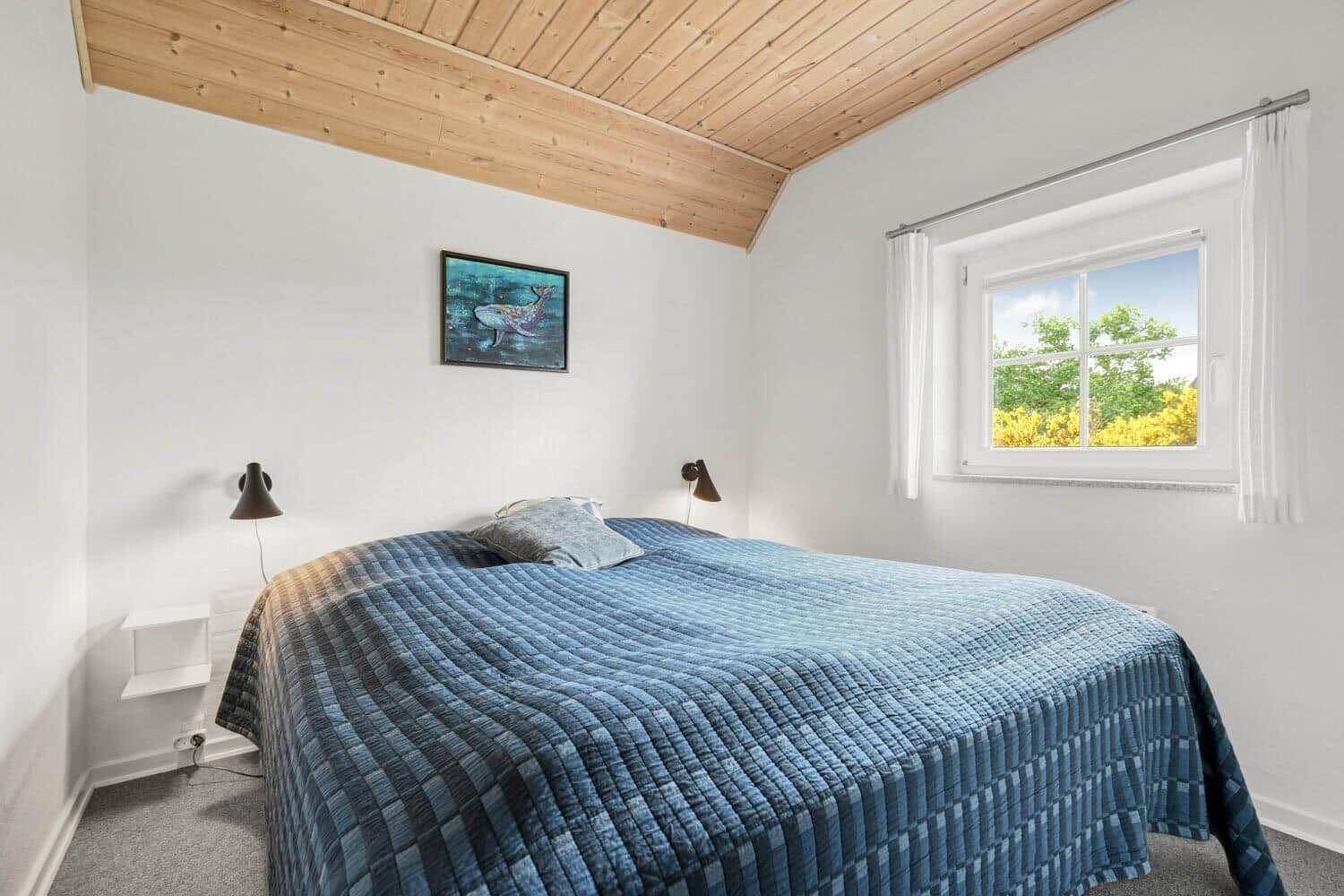 Schlafzimmer mit Bett, Wandbild und Fenster. Holzdecke und weiße Wände.