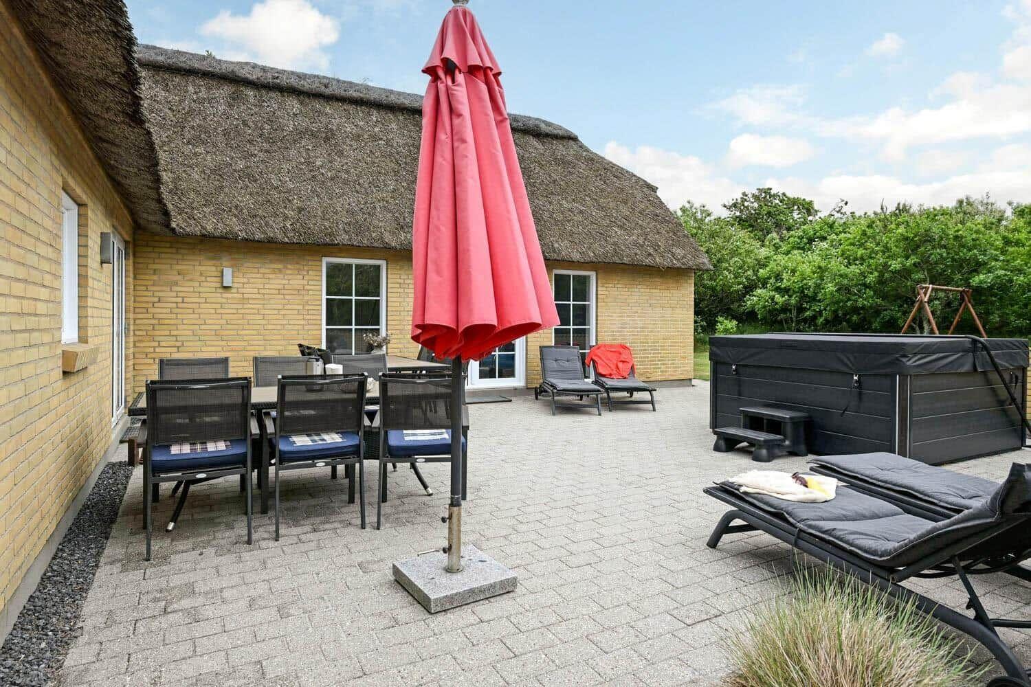 Terrasse mit Tisch, Stühlen, Sonnenschirm und Whirlpool. Hintergrund: Haus mit Strohdach.
